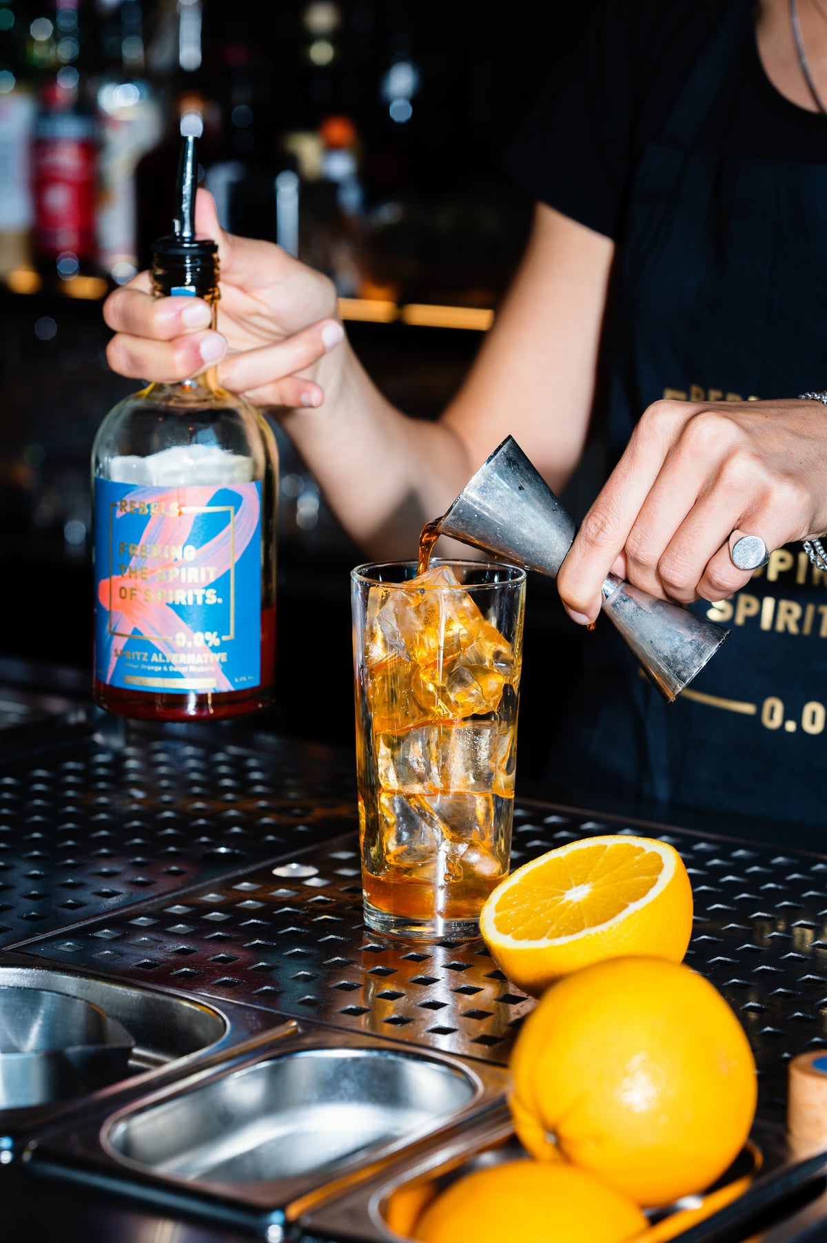 Bartender pouring a Rebels 0.0% Dolce Spritz into a glass with ice and oranges on a bar counter.