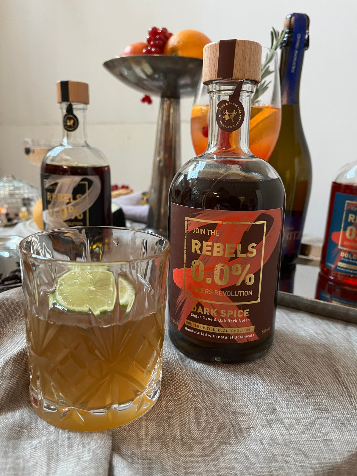 Bottle of Rebels 0.0% alcohol-free rum on a table with other bottles and a glass.